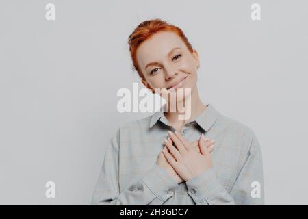 Junge reizende rothaarige Frau hält gefaltete Hände auf der Brust und fühlt sich dankbar und dankbar Stockfoto