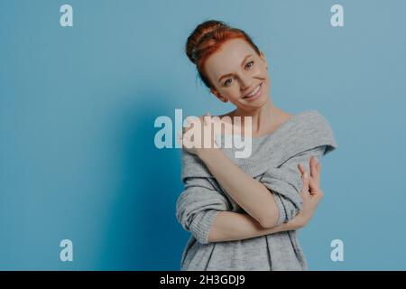 Fröhliche ruhige Ingwer-Frau, die Selbstliebe demonstriert, während sie sich gegen die blaue Wand posiert Stockfoto