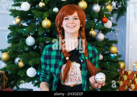 Charmante, lächelnde, rothaarige junge Frau im karierten Hemd vor dem Hintergrund eines weihnachtsbaums mit heller Beleuchtung in einem gemütlichen Wohnzimmer mit festlicher Stimmung. Stockfoto