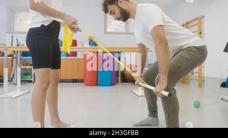 Paar während der Rehabilitationstherapie in der Klinik. Mann macht ein Ganzkörpertraining mit dem medizinischen Riegel und den Widerstandsbändern. Stockfoto