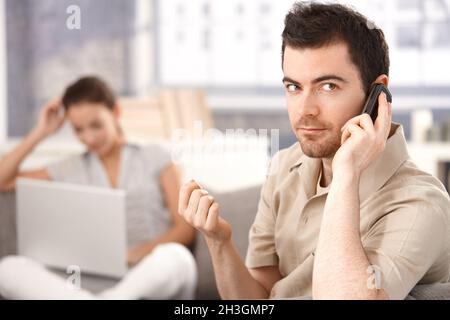 Junger Mann mit Handy zu Hause Frau im Hintergrund Stockfoto