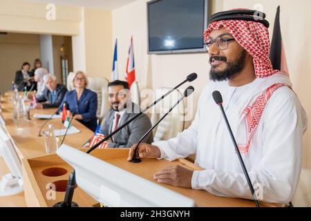 Lächelnder muslimischer Politiker in Ghutrah und Agal, der auf dem Podium steht und auf der internationalen Konferenz öffentlich mit Schleimphon spricht Stockfoto