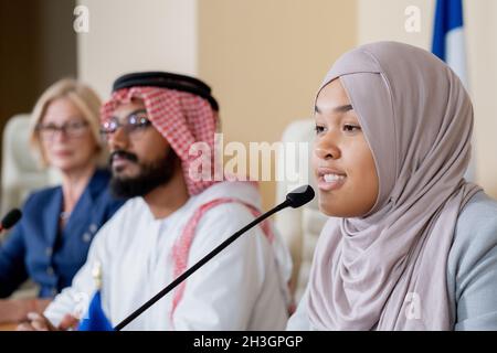 Attraktive junge muslimische Frau im Hijab, die während der Konferenzdiskussion ins Mikrofon spricht Stockfoto