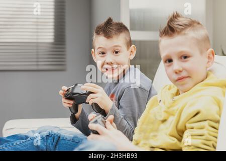 Schelmisch charismatischer Junge, der ein zotterisches Grinsen gibt, während er mit seinem Bruder Videospiele spielt und begeistert auf die Kamera blickt Stockfoto