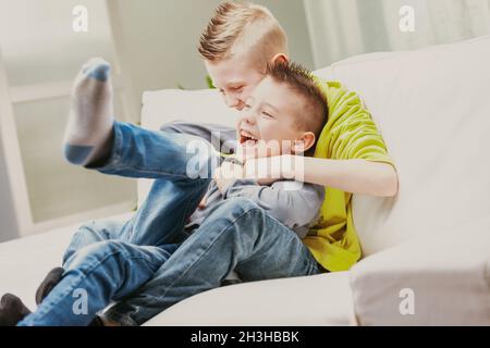 Zwei verschmitzte junge Brüder, die lachen, als sie auf der Couch im Wohnzimmer in einem hochaufragenden offenen Porträt toben Stockfoto