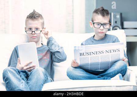 Parodie von zwei jungen Männern, die eine Brille tragen und sich als Geschäftsleute ausgeben, während sie Seite an Seite auf einem Sofa sitzen und die Business Times lesen und ein Tablet benutzen Stockfoto