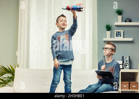 Zwei niedliche junge Jungs spielen zu Hause zusammen, einer mit Tablet und einer fliegenden Spielzeugrakete in der Luft Stockfoto