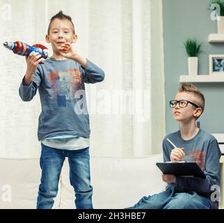 Zwei niedliche junge Jungs mit mohikanischem Spitzhaar spielen in einem Wohnzimmer mit einer Spielzeugrakete und konzentrieren sich auf einen Jungen, der vor der Kamera grinsend steht Stockfoto