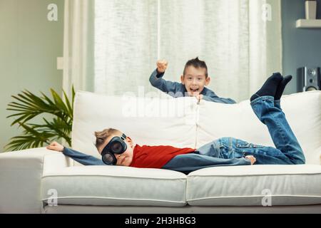 Zwei verschmitzte junge Brüder, die Spaß am Spielen auf einem Sofa haben, wobei einer von hinten schreit, während der zweite mit einer Brille auf dem Bauch liegt Stockfoto