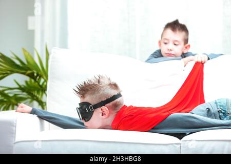 Kleiner Junge mit niedlicher, spiky Frisur, der Super Hero spielt, während er mit seinem roten Umhang durch die Luft fliegt, während er auf einem Sofa liegt Stockfoto