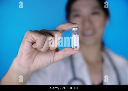 Eine attraktive Ärztin, die einen weißen Mantel mit einem Stethoskop um ihre Schulter trägt und eine Glasfläschchen mit Impfstoffflasche mit leerem weißen Etikett, Smili, hält Stockfoto