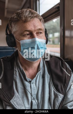 Porträt eines ernsthaften erwachsenen Mannes, der Kopfhörer mit einer medizinischen Schutzmaske auf seinem Gesicht in öffentlichen Verkehrsmitteln in der Nähe des Fensters trägt. Vorbeugende Maßnahme. Stockfoto