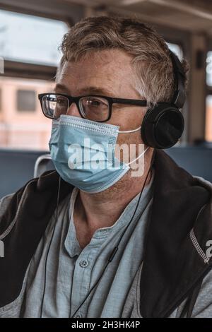 Nahaufnahme eines erwachsenen Mannes, der Kopfhörer und Brille mit einer medizinischen Schutzmaske auf seinem Gesicht in öffentlichen Verkehrsmitteln trägt. Soziale Distanz. Stockfoto