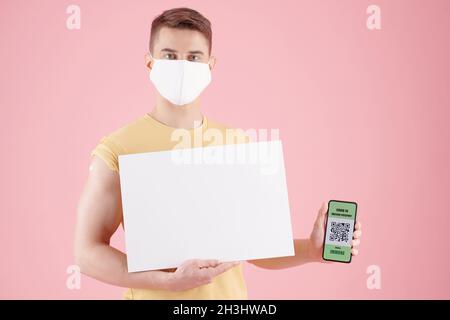 Seriöser junger Mann in Schutzmaske mit schwarzem Plakat und Smartphone mit QR-Code und Impfpass Stockfoto