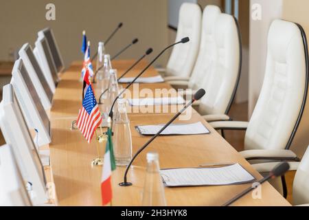 Monitore gegen Bürostühle im Konferenzraum: Klemmbretter, Flaschen, Mikrofone und Nationalflaggen auf Holztisch Stockfoto