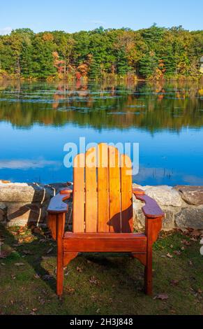 Adirondack-Stuhl vor einem See, an einer Steinmauer. Mischwald mit Bäumen in Herbstfarben im Hintergrund. Moore State Park, Paxton, MA Stockfoto