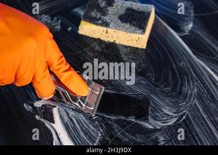 Nahaufnahme eines orangefarbenen Handschuhs mit einem Metallschaber, der ein Kochfeld reinigt. Selektiver Fokus. Ein Schwamm liegt im Hintergrund. Das Konzept der Hausreinigung ein Stockfoto