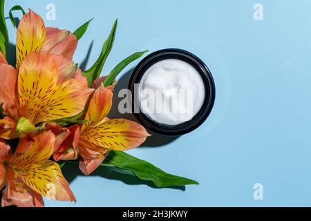 Offene Kosmetikgläser mit pflegender oder feuchtigkeitsspendender Creme auf blauem Hintergrund und Alstroemeria-Blume mit Kopierfläche. Stockfoto