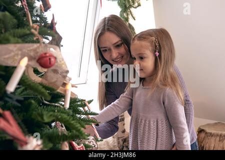 Mutter und Tochter schmücken den Weihnachtsbaum. Portrait liebevolle Familie in schönen holdiay DIY handgemachte Wohndekoration. Gemütliches Zimmer mit Kerzenlicht eingerichtet Stockfoto