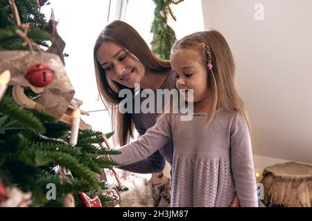 Mutter und Tochter schmücken den Weihnachtsbaum. Portrait liebevolle Familie in schönen holdiay DIY handgemachte Wohndekoration. Gemütliches Zimmer mit Kerzenlicht eingerichtet Stockfoto