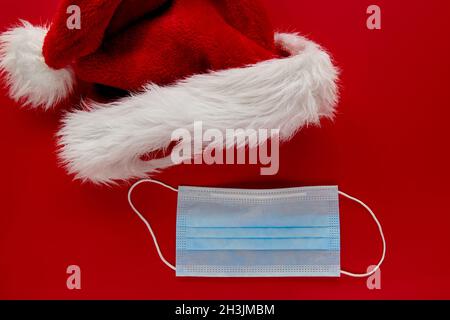 Festlicher Weihnachtsmann-Hut mit Coronavirus, Gesichtsmaske und Verzierungen auf rotem Hintergrund. Flat Lay, Draufsicht weihnachtsfeiertage Komposition. Wallp. Neujahr Stockfoto