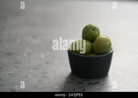 Große grüne Oliven in schwarzer Schale auf Beton Hintergrund, flacher Fokus Stockfoto