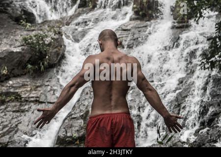 Rückansicht eines muskulösen Mannes, der mit erhobenen Armen in der Nähe des Wasserfalls steht Stockfoto