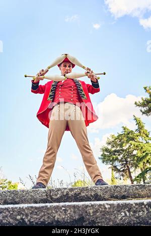 Bärtiger erwachsener Mann in rotem Anzug, der mehrere Keulen hält und die Kamera anschaut, während er während der Show im Park gegen den wolkigen blauen Himmel steht Stockfoto