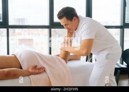 Verkürzte Seitenansicht des professionellen männlichen Masseurs mit starken Händen, die nach dem Sport-Training die Wadenmuskulatur am Unterschenkel des Muskelathleten massieren Stockfoto