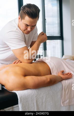 Vertikale Aufnahme eines männlichen Masseurs, der den Rücken eines muskulösen Sportlers massiert und auf dem Massageliegen mit dem Ellbogen auf dem Hintergrund des Fensters liegt. Stockfoto