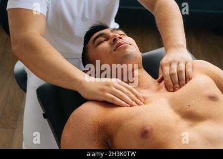 High-Angle-Ansicht des professionellen männlichen Masseurs mit starken Händen, die die Schulter- und Brustmuskeln des Muskelathleten Mann bei einer Spa-Behandlung massieren, Stockfoto