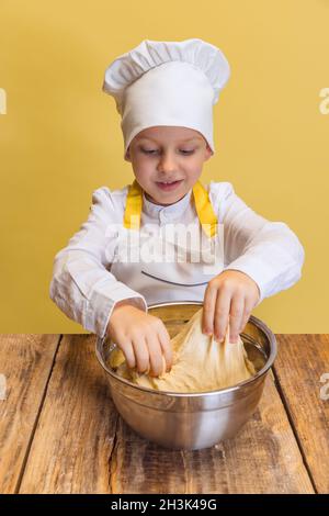 Netter lächelnder Junge in weißem Koch, Kochjacke und Hut knetet Teig isoliert auf gelbem Studiohintergrund. Stockfoto