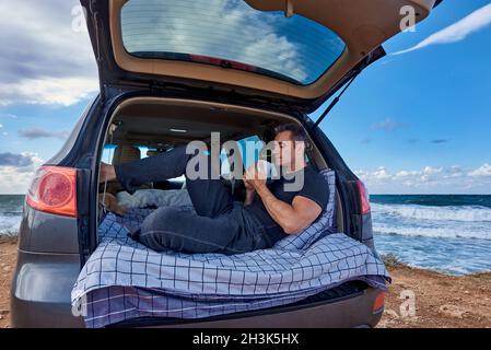 Ein Mann trinkt Kaffee, während er auf einer Fahrt am Meer in seinem Kofferraum sitzt. Stockfoto