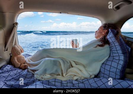 Eine junge Frau, die mit offenem Lastwagen in einem Auto sitzt, bedeckt mit einer gemütlichen Decke, während sie den Blick auf das Meer betrachtet. Stockfoto