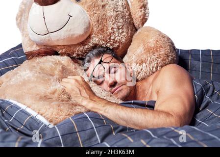 Ein junger Mann schläft in einer Umarmung mit einem Teddybär im Bett auf weißem Hintergrund. Stockfoto