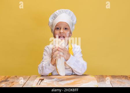 Ein süßer Junge trägt einen weißen Koch, eine Chefjacke und einen Hut und knetet Teig isoliert auf gelbem Studiohintergrund. Stockfoto