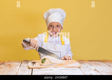 Comic-Porträt des kleinen Jungen trägt in weißen Koch, Chefjacke und Hut knetet Teig isoliert auf gelben Studio-Hintergrund. Stockfoto