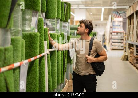 Nachdenkliche Casual Kerl im Geschäft mit Hauswaren Auswahl Kunstrasen in Rollen auf dem Display Stockfoto