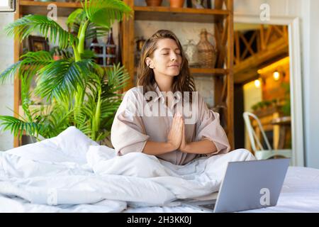 Junge Erwachsene Frau meditiert im Bett vor dem Laptop-Monitor - Konzept der Wiederherstellung von Kraft und geistiger Energie durch Yoga-Praxis. Stockfoto