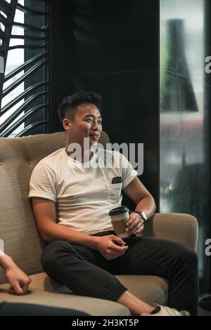 Asiatischer junger, gutaussehender Mann, der im Café sitzt, Kaffee trinkt, redet und lächelt.hübscher junger Mann, Leben der Millennials, Lebensstil in der Großstadt Stockfoto