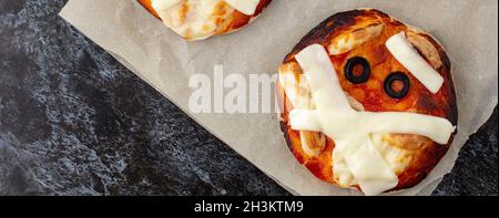 Banner von Mini Pizza als Mama für Kinder mit Käse, Oliven und Ketchup. Witziges verrücktes Halloween Essen für Kinder auf dunklem Hintergrund Stockfoto