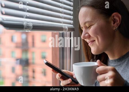 Fröhliche lächelnde Frau mit Tasse, die das Telefon neben dem Fenster mit Jalousien benutzte Stockfoto