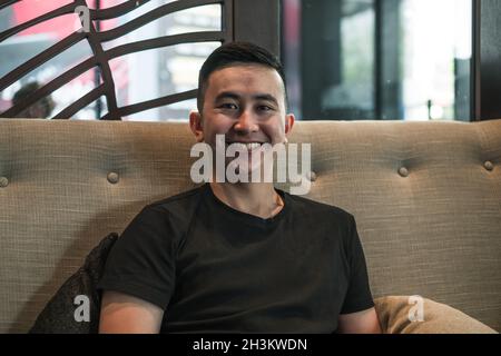 Porträt eines jungen, hübschen asiatischen Mannes in schwarzem T-Shirt, der im Café sitzt und lächelt. Schönes Lächeln, glückliche Person, Millennials Leben, Lebensstil Stockfoto