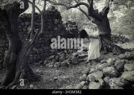 Gesichtslose, geisterhafte asiatische Frau, die in einer verlassenen, verdrehten Olivenhainplantage in der Nähe einer alten christlichen Kapelle spazierengeht. Stockfoto