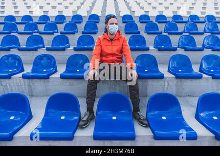 Fußballtribünen mit roten und blauen Plastikstühlen. Fußballfan mit Schutzmaske im Stadion. Sportwettkämpfe während Quarantäne und Lockdo Stockfoto