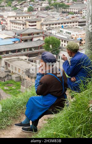 Tibetische Männer sitzen, verweilen die Zeit, indem sie eine Zigarette rauchen und entspannen sich auf dem Hügel mit Blick auf die alte, von Mauern umgebene chinesische Stadt mit Song Pain, Nordsichuan, China. (125) Stockfoto
