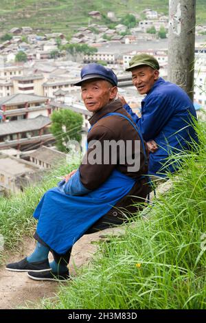 Tibetische Männer sitzen, verweilen die Zeit, indem sie eine Zigarette rauchen und entspannen sich auf dem Hügel mit Blick auf die alte, von Mauern umgebene chinesische Stadt mit Song Pain, Nordsichuan, China. (125) Stockfoto