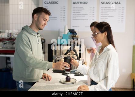 Mann in legerer Kleidung, der asiatischen Gästen im Kaffeehaus heiße Getränke und Desserts serviert Stockfoto