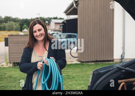 Porträt einer lächelnden Frau, die das Ladekabel eines Elektroautos an der Station hält Stockfoto