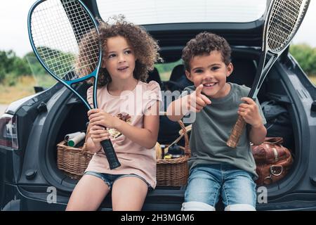 Junge hält Tennisschläger, während sie auf die Schwester zeigt, die im Kofferraum sitzt Stockfoto